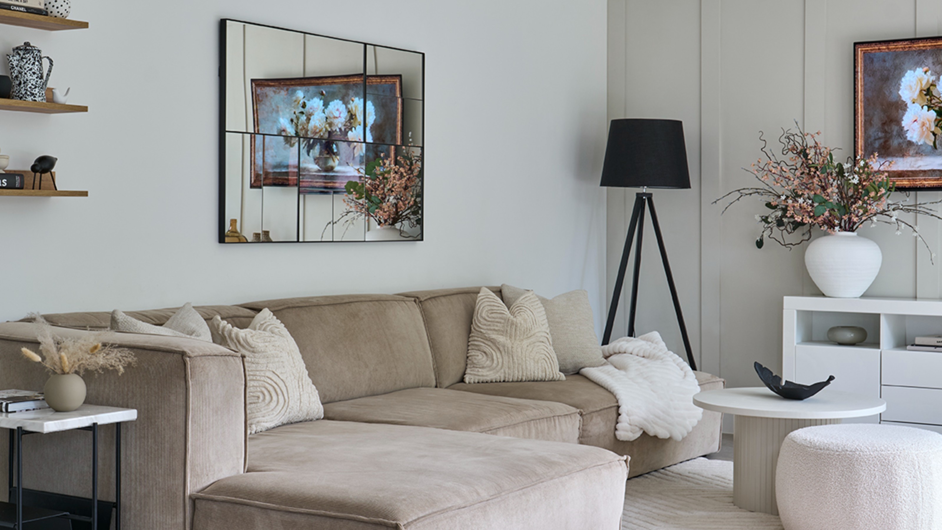 A neutral living room with a beige modular sofa and a rectangular windowpane-style mirror above it reflecting an artwork-displaying TV