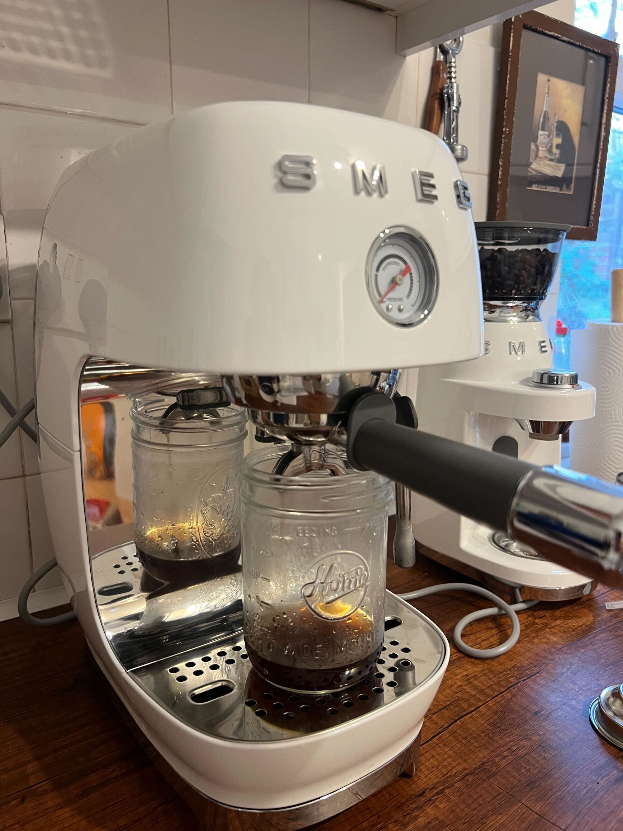 Image of a white Smeg cold brew machine brewing cold brew into a mason jar on a wooden countertop with white tile backsplash