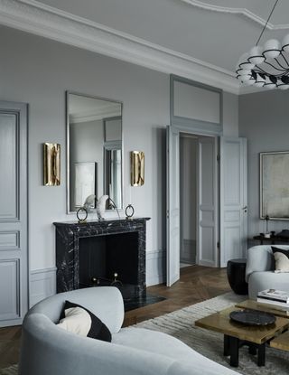 ornate light blue living room with marble fireplace, a mirror hung above with two wall sconces on eithe side, two blue sofas facing each other with a gold coffee table in the middle