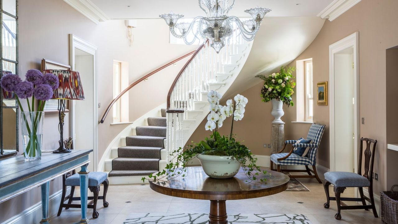 Entryway with pale pink walls, round wooden table with orchid, chandelier, mirror on wall and blue console table underneath