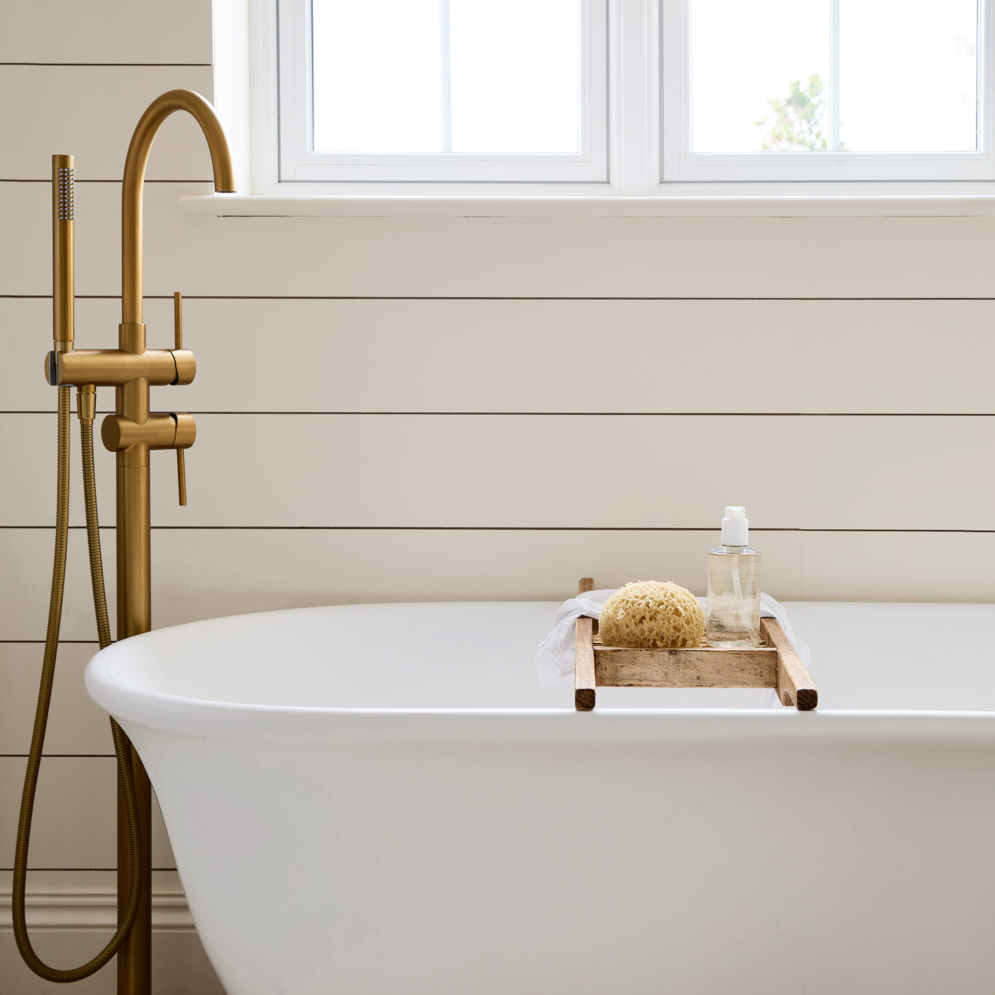 close up of a neutral bathroom with horizontal wall panelling, a large white freestanding bath tub and brass fixtures