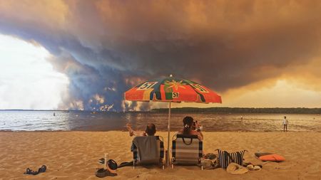TOPSHOT - Beach goers rest on the beach of Moutchic on the shore of Lake Lacanau (Etang de Lacanau) in Lacanau , southwestern France on July 24, 2026, as clouds of smoke rise into the sky from the Gironde wildfire. The fire raging in the northern part of the Arcachon Basin in Gironde, southwest France, has swept through more than 3,400 hectares, according to the latest figures released by the prefecture. More than 20,000 residents and tourists have been evacuated from this part already devastated by flames in 2022. Scientists have long warned that climate change caused by human use of fossil fuels will make periods of drought more intense and longer-lasting, creating the ideal conditions for wildfires. (Photo by Maximilien LAMY / AFP via Getty Images)