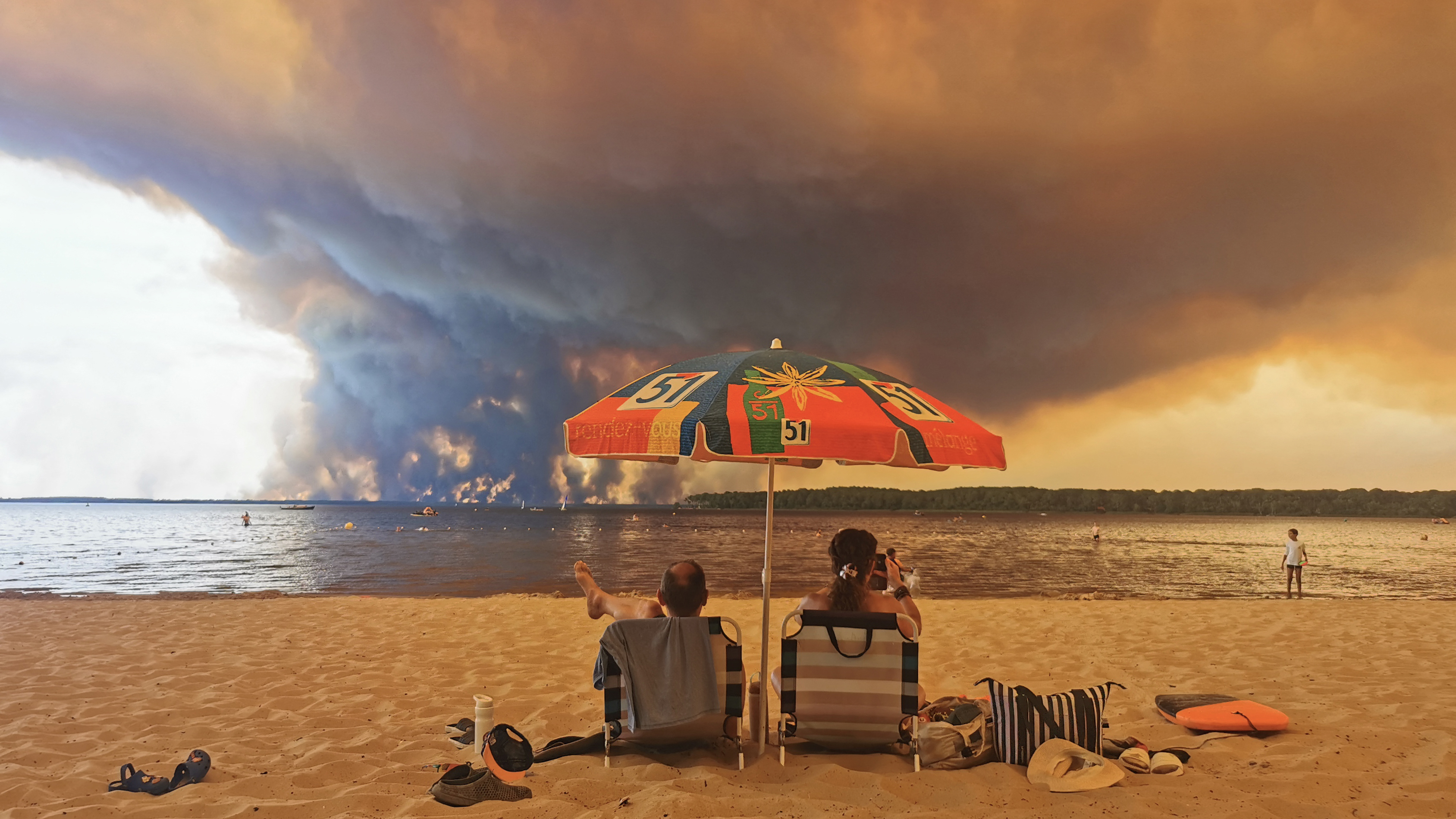 TOPSHOT - Beach goers rest on the beach of Moutchic on the shore of Lake Lacanau (Etang de Lacanau) in Lacanau , southwestern France on July 24, 2026, as clouds of smoke rise into the sky from the Gironde wildfire. The fire raging in the northern part of the Arcachon Basin in Gironde, southwest France, has swept through more than 3,400 hectares, according to the latest figures released by the prefecture. More than 20,000 residents and tourists have been evacuated from this part already devastated by flames in 2022. Scientists have long warned that climate change caused by human use of fossil fuels will make periods of drought more intense and longer-lasting, creating the ideal conditions for wildfires. (Photo by Maximilien LAMY / AFP via Getty Images)