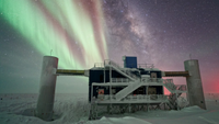 The NSF IceCube Lab is seen under a starry, night sky, with the Milky Way appearing next to striking auroras in the background
