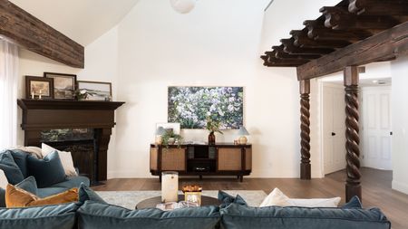 Large living room space with blue sofa, wooden beams, a retro media unit, a piano, and framed artwork on white walls.