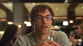 Robert Pattinson wears glasses and a grey shirt while sitting at a restaurant and holding a wine glass in The Drama. 