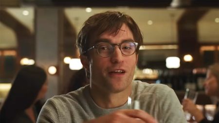 Robert Pattinson wears glasses and a grey shirt while sitting at a restaurant and holding a wine glass in The Drama. 
