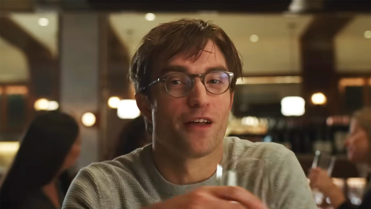 Robert Pattinson wears glasses and a grey shirt while sitting at a restaurant and holding a wine glass in The Drama. 