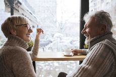A man and woman wearing sweaters talk at a cafe with a snowy background behind the windows.