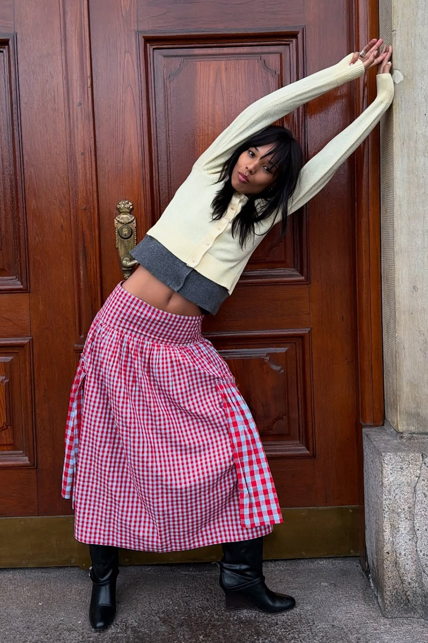 a woman wearing a butter yellow cardigan, red gingham skirt, and black boots