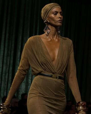 Saint Laurent model wearing a tan cap with a matching low-cut blouse, belted skirt, and oversize earrings.