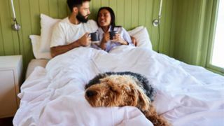 A couple lie in bed with their dog sleeping at the foot of the bed