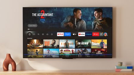 The Amazon Fire TV Omni QLED (2025 edition) displaying the Fire TV smart platform home screen in a living room setting
