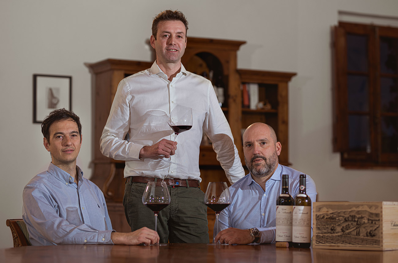 Three men, two sitting behind a table and one standing. All are holding a glass of red wine, and there are two bottles on the table