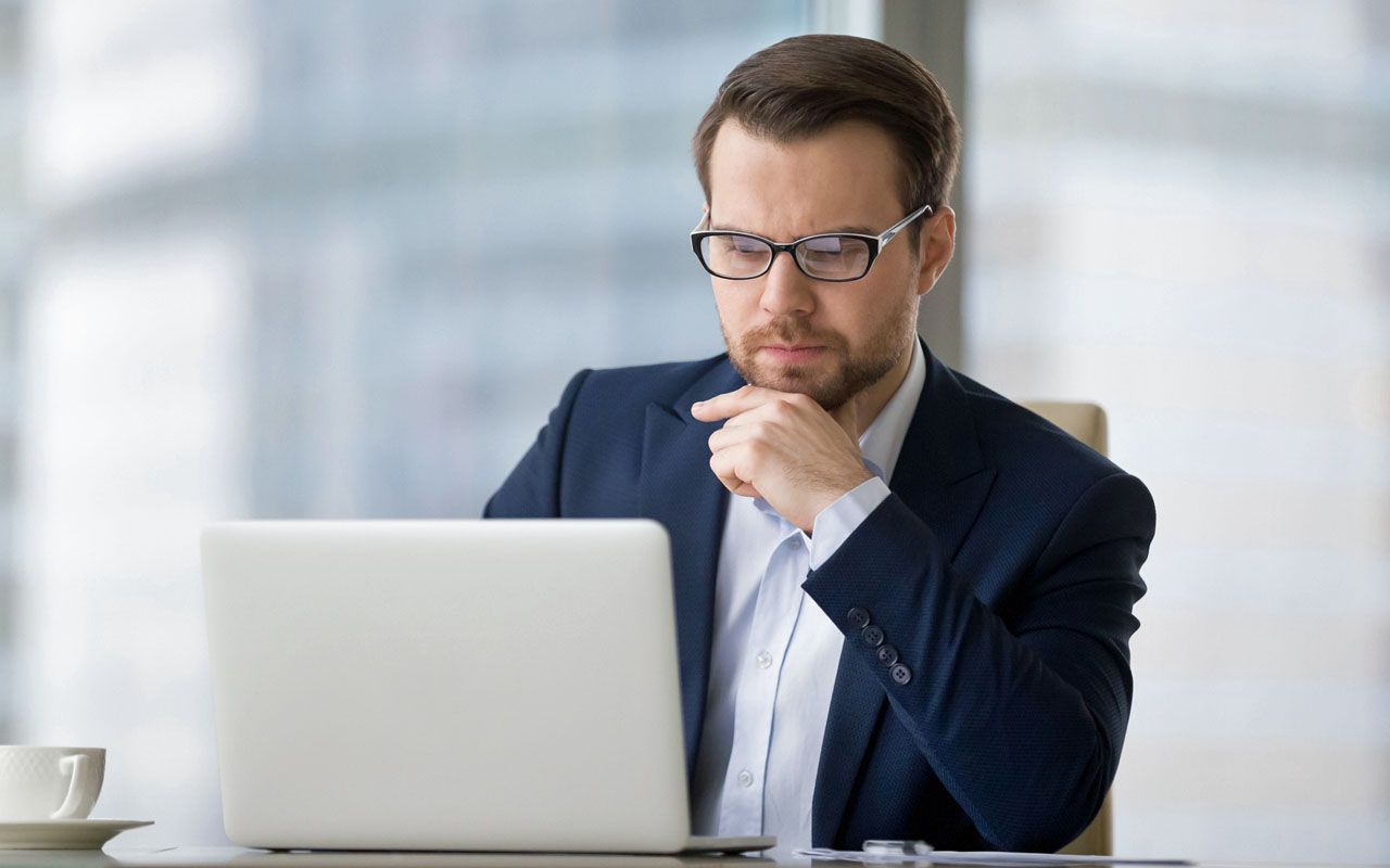 Caucasian man sitting at the table near laptop in the office and looking at the screen. Serious and pensive boss or employee working at his workplace and feeling interest or analyzing results