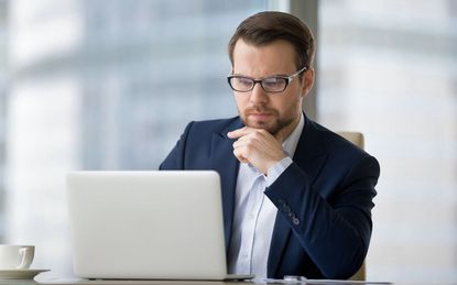 Caucasian man sitting at the table near laptop in the office and looking at the screen. Serious and pensive boss or employee working at his workplace and feeling interest or analyzing results