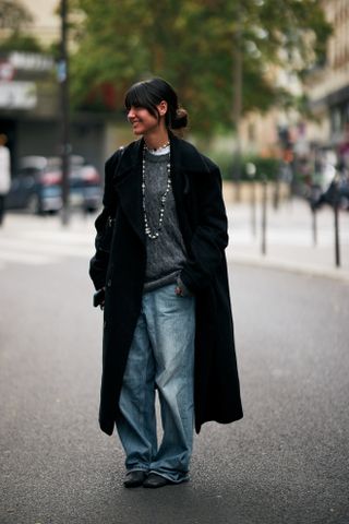 A woman wearing a gray sweater, white long sleeve shirt, blue jeans, black boots, black coat, and silver necklace