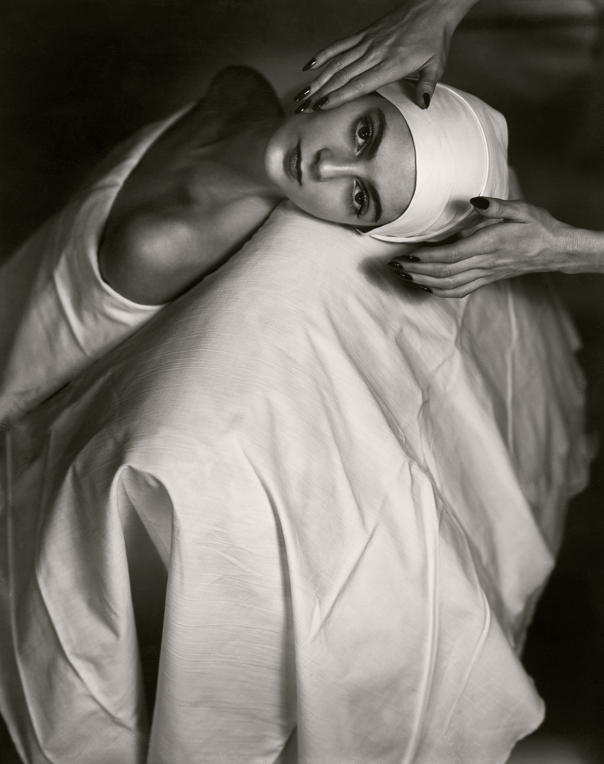 A black-and-white close-up shows a model's face resting on a draped white cloth as two hands gently frame her head.
