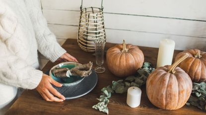 Someone arranging a bown of linenes on a wooden table covered in fall pumpkins and foliage