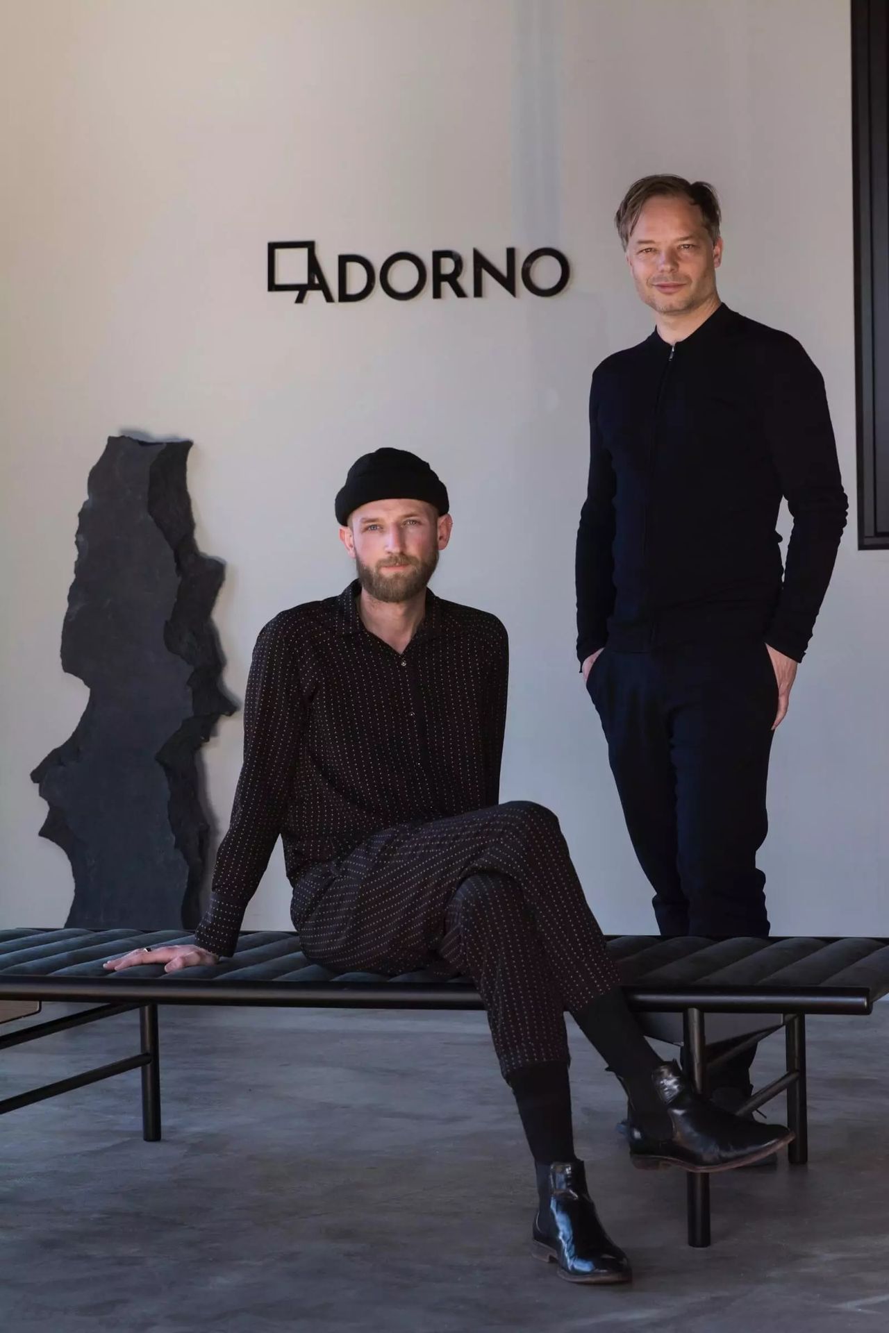 Two men dressed in black sit in a dimly lit gallery.