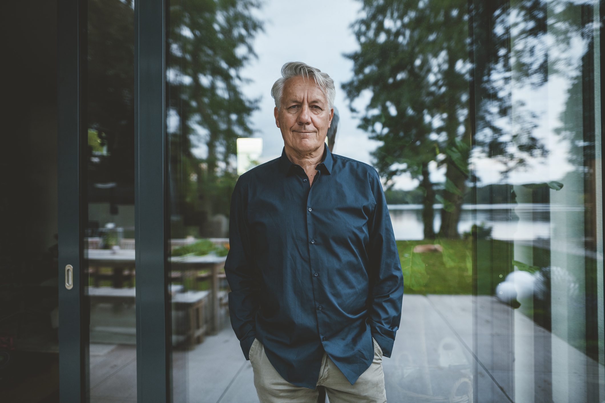 Senior man with hands in pockets standing in front of glass wall.