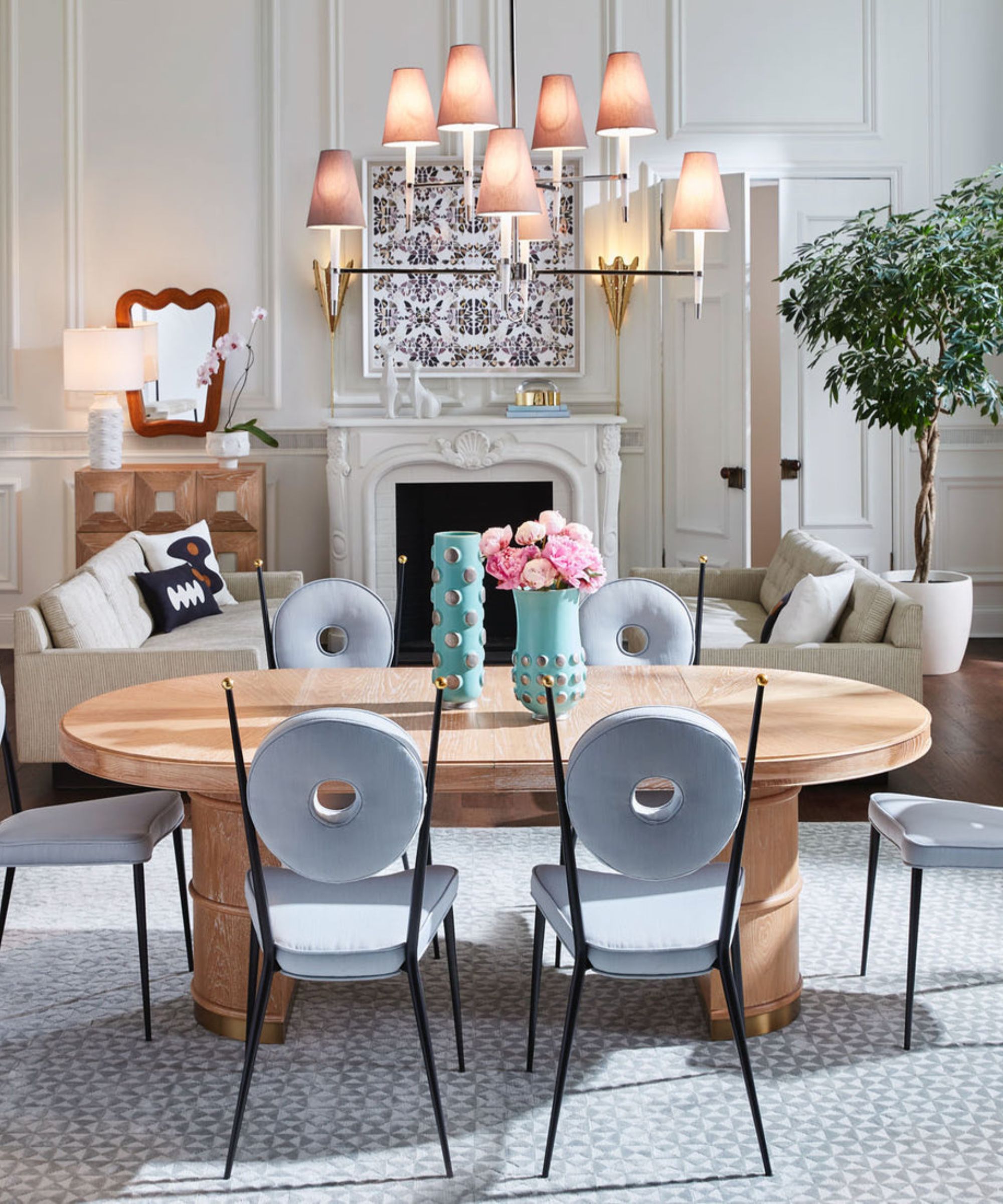 A sophisticated dining room showing a light wood dining table with "donut" back chairs, turquoise studded vases, and a white fireplace in the background