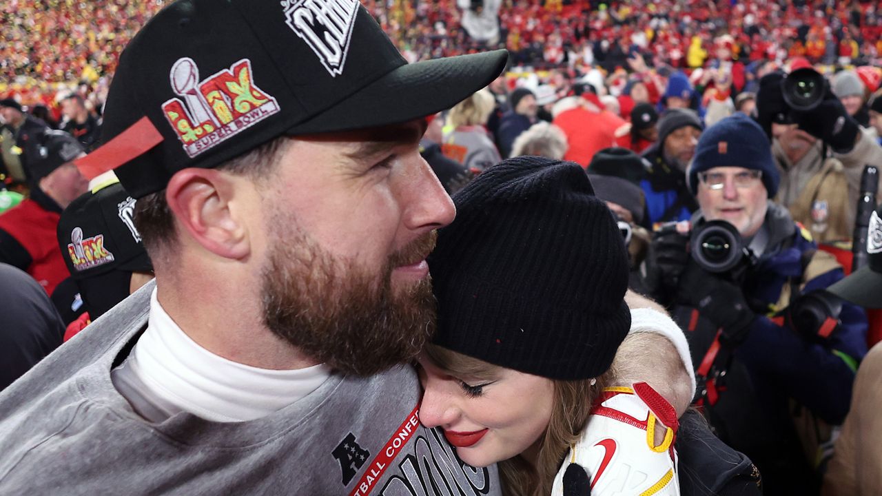 Travis Kelce wearing a black baseball cap hugging Taylor Swift, wearing a black winter hat, on the field after the AFC Championship game 