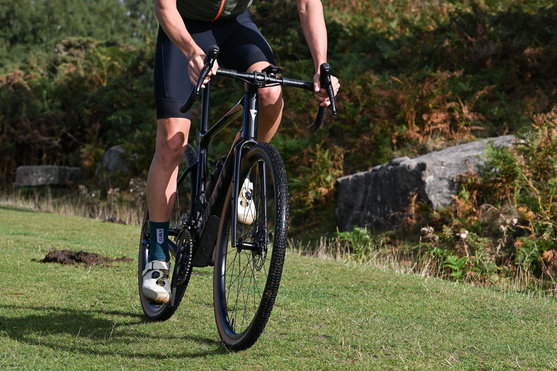man riding a black gravel bike wearing black shorts, blue socks and beige SPD shoes riding on a grassy track