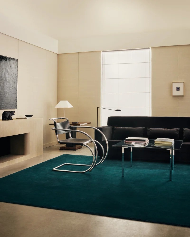 A modern living room with off-white walls, a dark teal rug, and black and chrome modern furniture.
