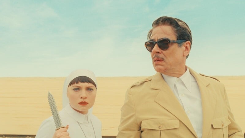 Mia Threapleton holds a knife and Benicio Del Toro wears sunglasses in a desert in The Phoenician Scheme