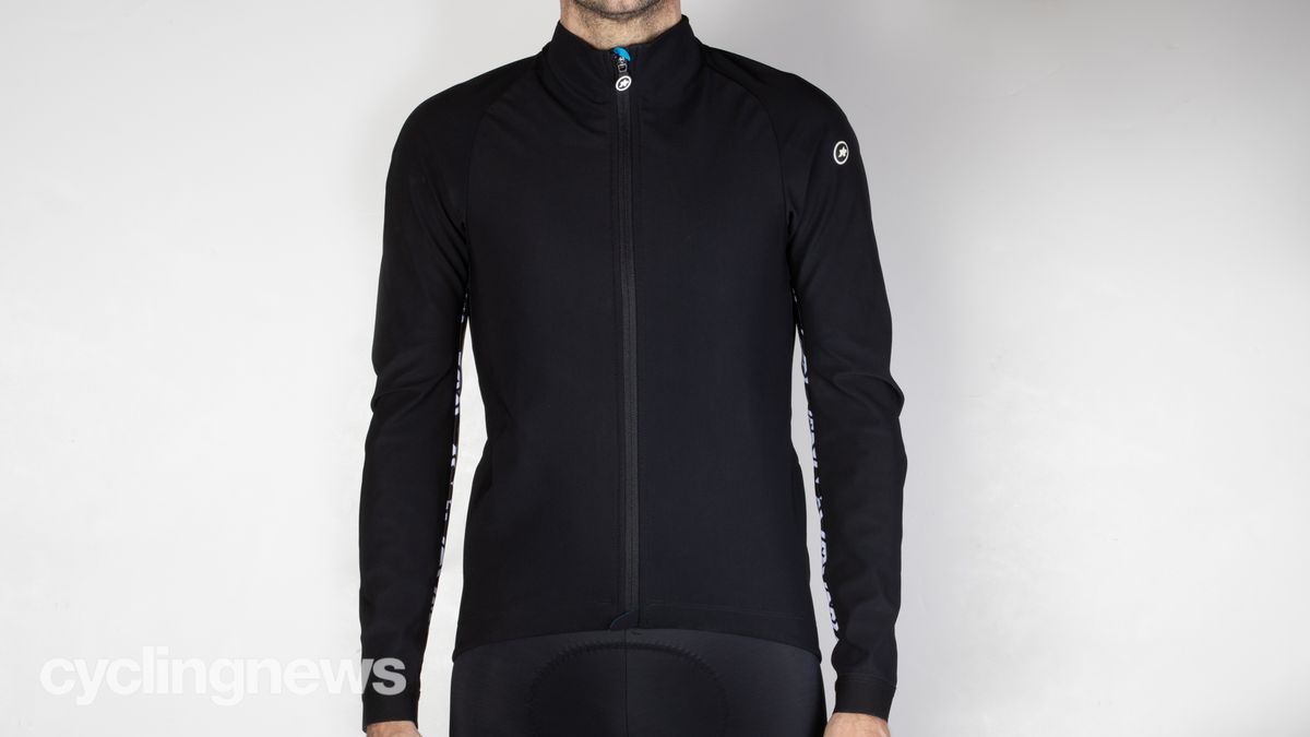 A white man in a black winter cycling jacket stands against a grey wall
