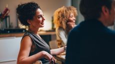 Woman looking out at table of friends, smiling at conversation with glass in hand sitting in kitchen