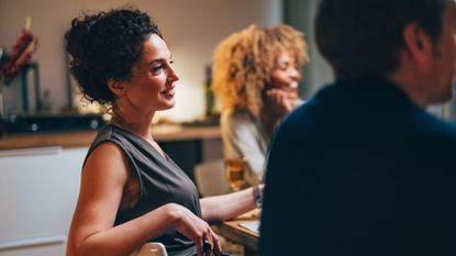 Woman looking out at table of friends, smiling at conversation with glass in hand sitting in kitchen