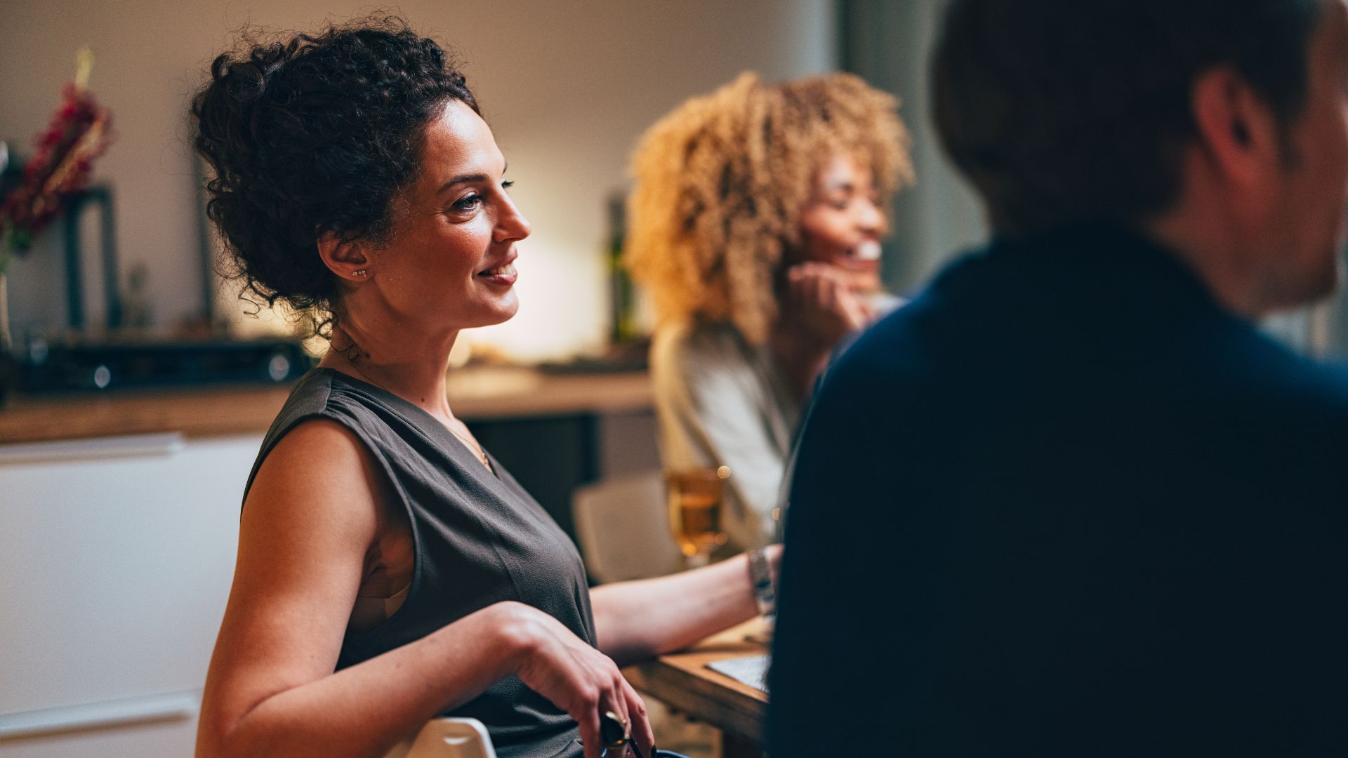 Woman looking out at table of friends, smiling at conversation with glass in hand sitting in kitchen