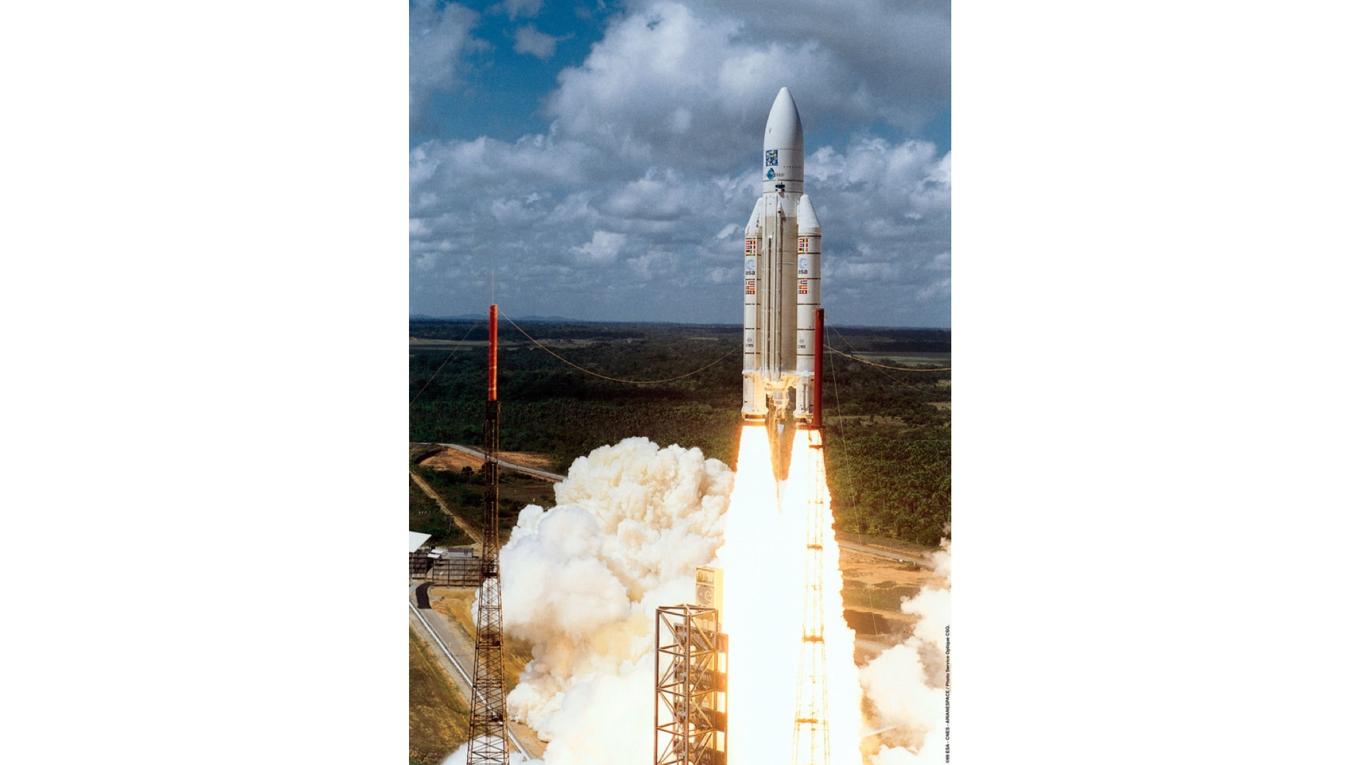 An ariane 5 rocket lifts off from its launchpad against the backdrop of green forest and blue skies.