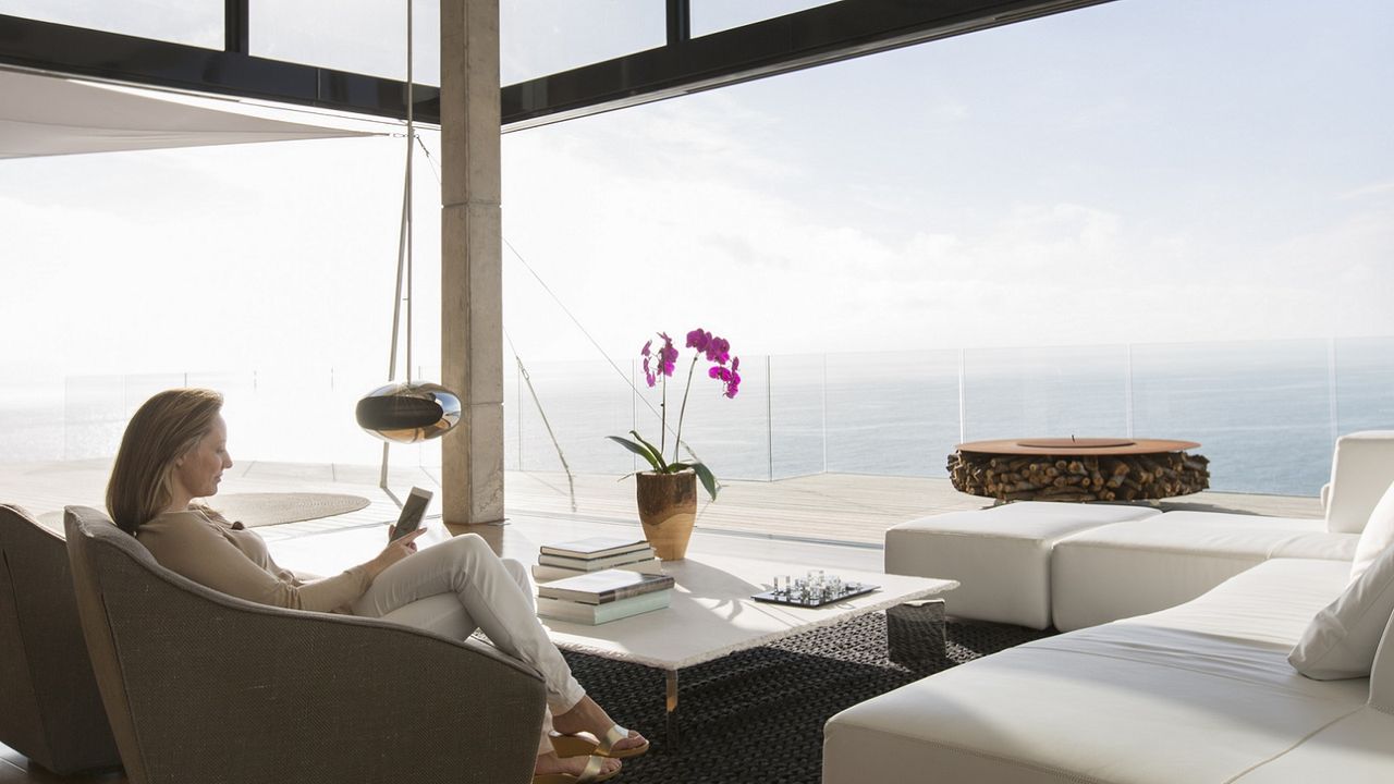 An older woman sits on a couch in a luxury home looking out at the ocean.