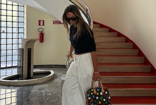 Hanna MW wearing a black t-shirt, white trousers, and a Louis Vuitton Speedy bag next to a red spiral staircase.