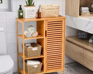 A wooden freestanding bathroom cabinet with rolled up towels in wicker baskets and accessories