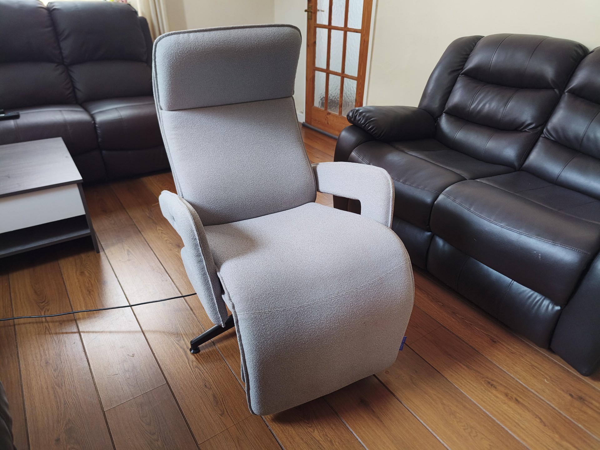 Three-quarter angled view of the upright grey fabric Neurosonic Serene recliner in a living room, flanked by two dark leather sofas.