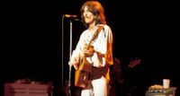 George Harrison wears all white and plays an acoustic guitar during his 1974 Dark Horse tour.