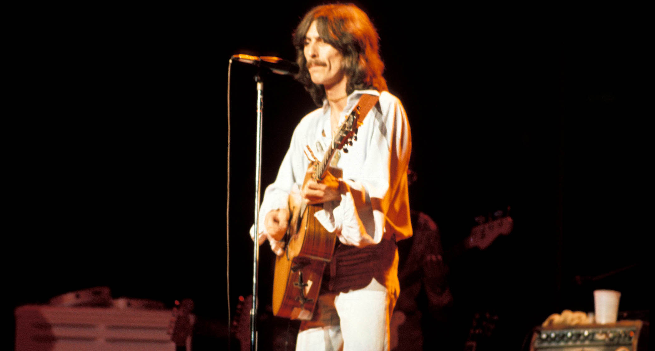 George Harrison wears all white and plays an acoustic guitar during his 1974 Dark Horse tour.