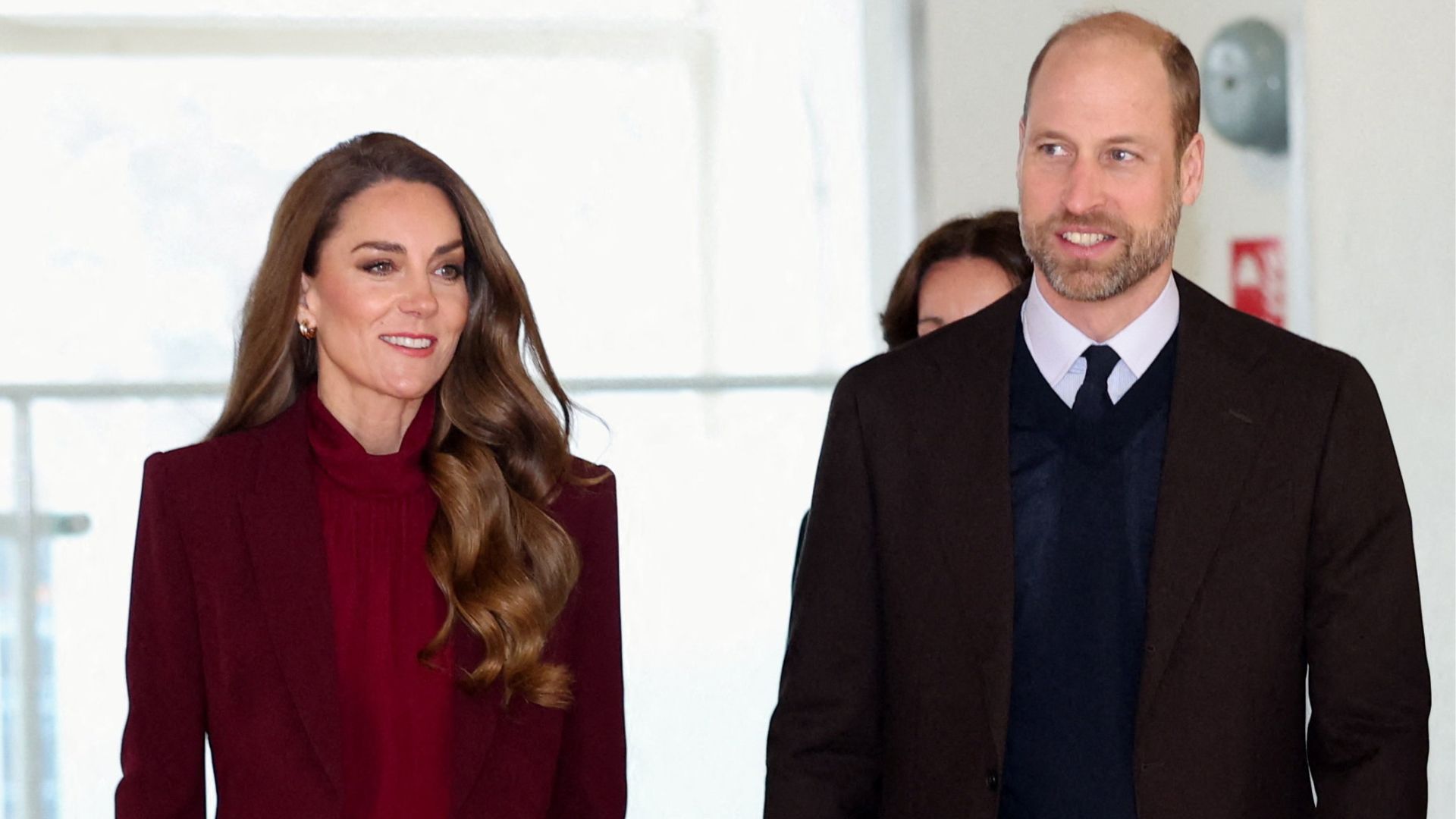 Kate Middleton walks next to Prince William wearing a burgundy suit with a brown bag and shoes.