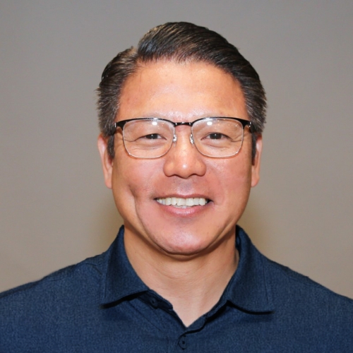 Image of a man with glasses in a dark blue collared shirt standing in front of a gray backgorund.