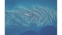 A bright blue and turquoise ocean has a center of waves and ripples of sand with white clouds overhead as seen from above. 