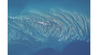 A bright blue and turquoise ocean has a center of waves and ripples of sand with white clouds overhead as seen from above. 