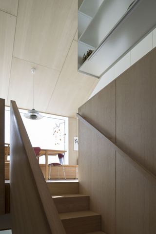 Interior view of the wooden steps leading to the main living area at the Oslo family house. There is a partial view of the living area featuring a wood panelled ceiling and walls, a dining table, red chairs, shelving and a pendant light