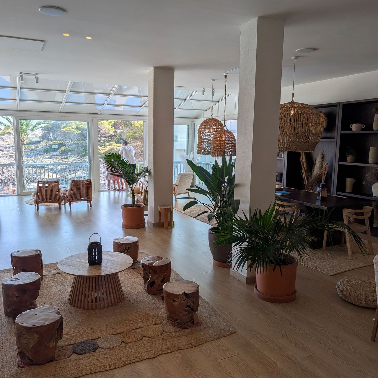 Open-plan living area with floor-to-ceiling windows, wooden stools, jute rugs and house plants