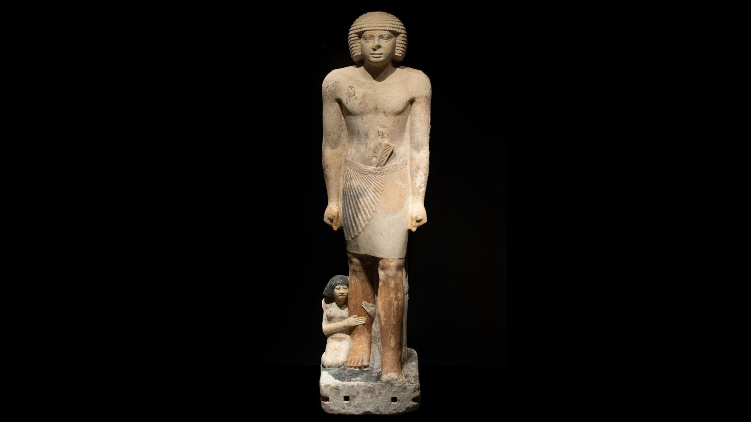 A limestone statue of a man standing tall and his tiny wife on her knees and holding onto his leg.