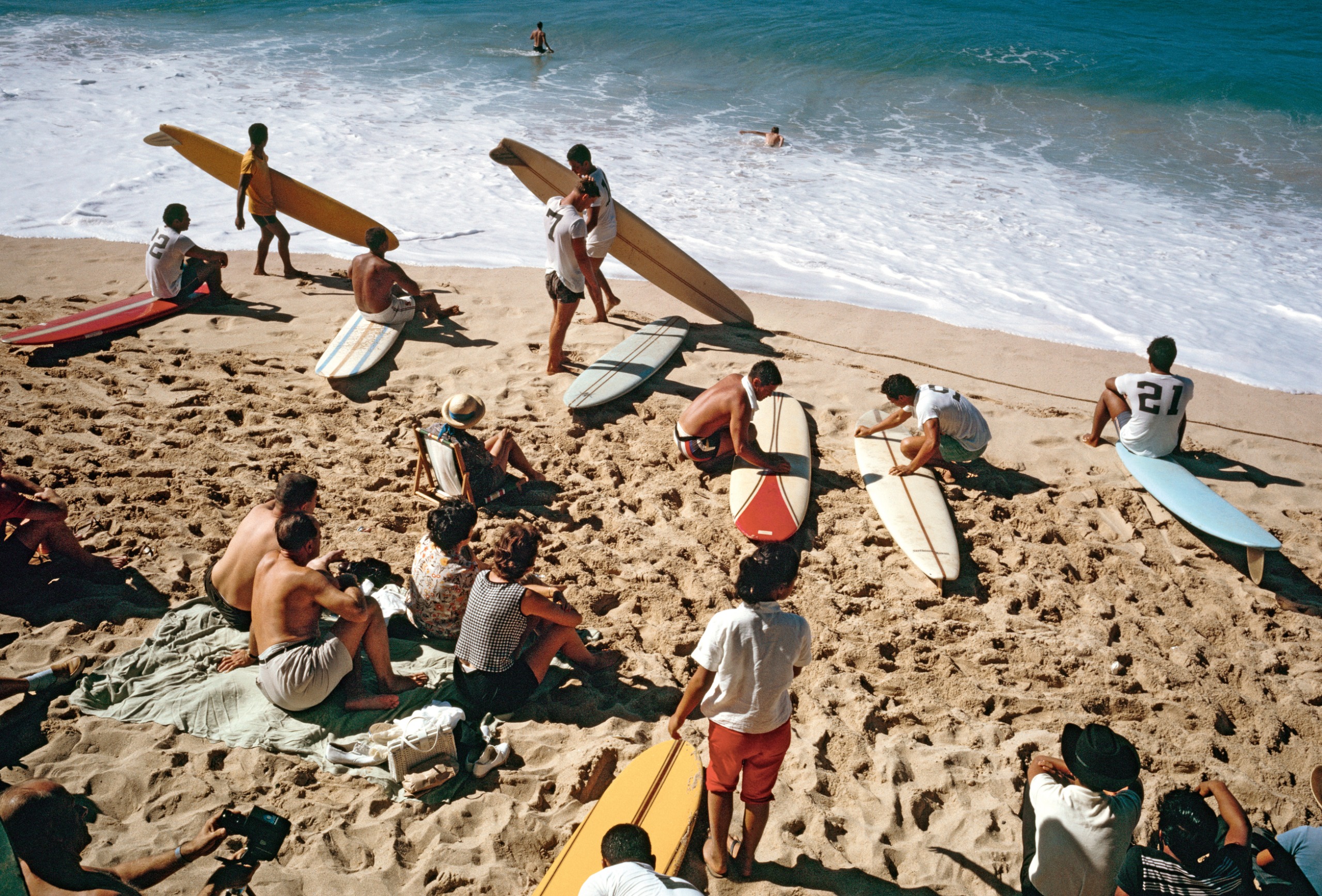 An overhead shot of surfers and sunbathers on Mākaha Beach, Hawaii, with longboards propped upright in the sand and two swimmers visible in the breaking waves.