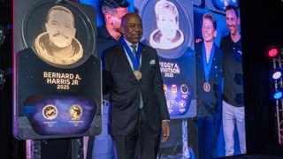 a Black man in a blue suit wearing a medal poses with a plaque depicting him and his astronaut mission patches. Behind him is a video screen showing the same with a woman.